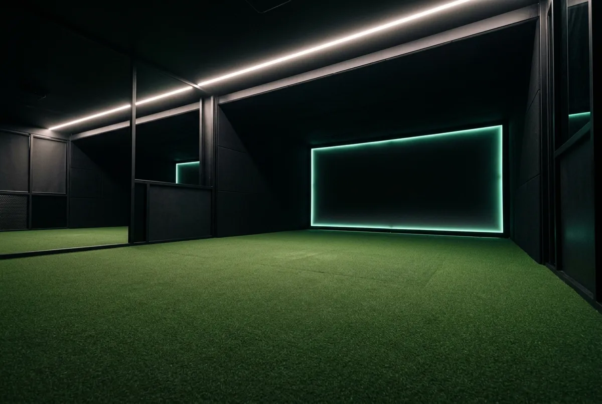 An indoor golf simulator bay at night — premium turf, overhead track lighting, dark walls — the SwingSpot interior.